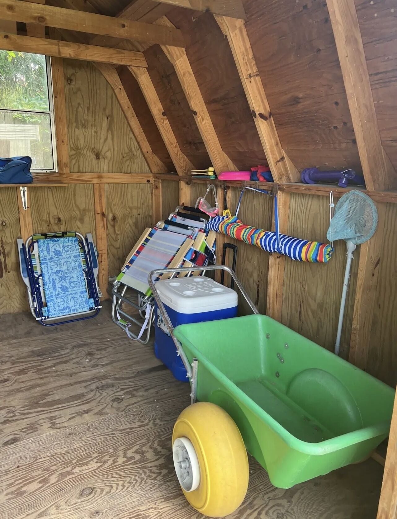A small, wooden attic room with a sloped ceiling, a green wheelbarrow, a blue cooler, and a colorful toy on a rack.