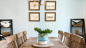 Una sala da pranzo con un lungo tavolo in legno, sei sedie in legno, un runner blu, un centrotavola con una pianta decorativa e quadri appesi alle pareti.