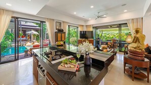 Une cuisine moderne avec un îlot central, un coin repas agrémenté d’une statue de Bouddha et une vue sur une piscine et la verdure environnante.