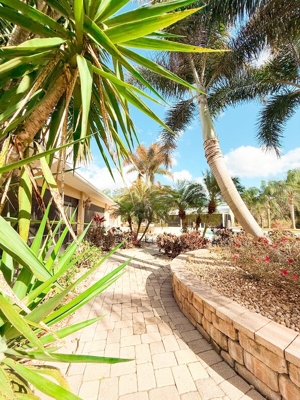 A paved pathway leading to a building with a stone wall and a variety of plants and trees.