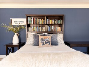A bedroom with a large bookshelf filled with books, a white vase with flowers, and a 'Game Day' pillow on the bed.