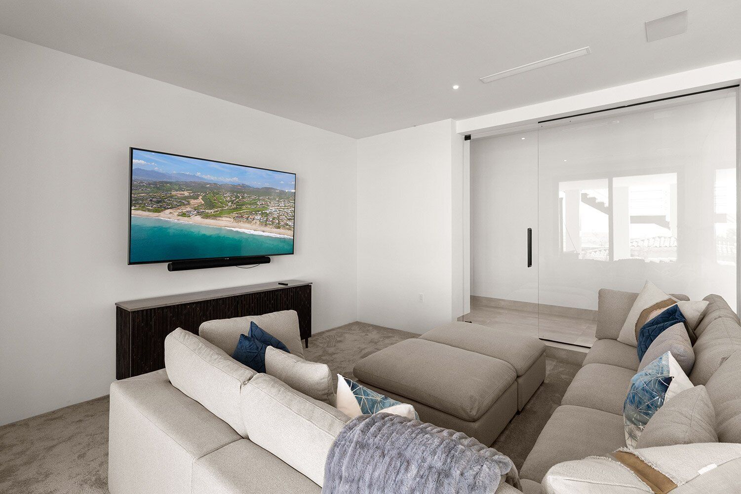 Ein modernes Wohnzimmer mit einem großen Flachbildfernseher an der Wand, einem Ecksofa mit vielen Kissen und einer Glastür, die auf einen Balkon führt.