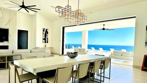 Un comedor moderno con una gran mesa y sillas, un ventilador de techo y vista al océano.