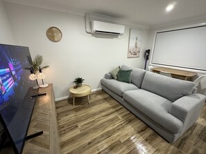 Un salon moderne avec un canapé gris, une petite table d’appoint ronde en bois, un téléviseur à écran plat et un climatiseur fixé au mur.