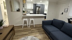 A modern living room with a blue sofa, a small coffee table, and a wooden side table with a plant. A kitchen with white cabinets and a gray countertop is visible in the background.