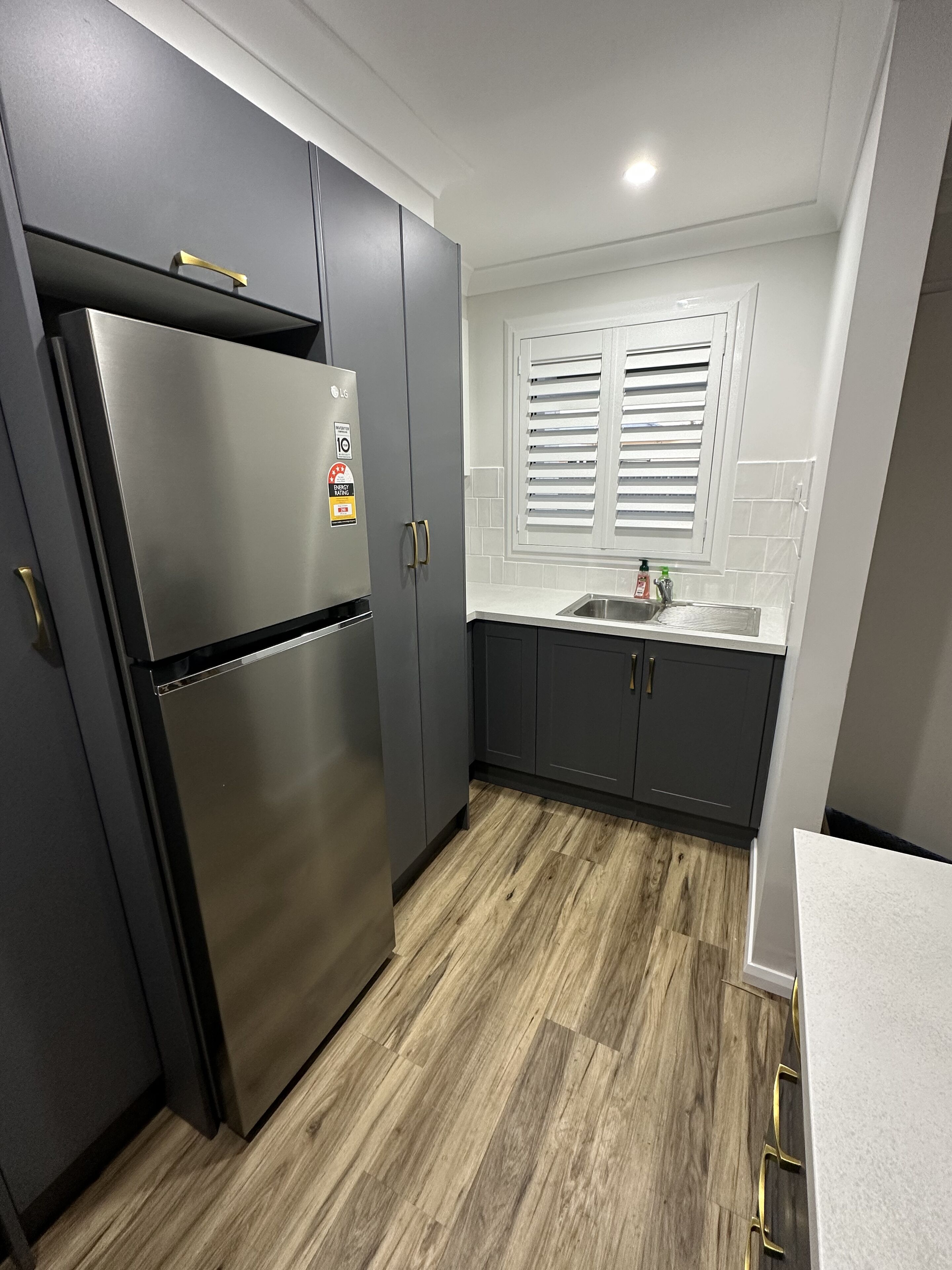 A modern kitchen with dark cabinets, a stainless steel refrigerator, a window with blinds, and a wooden floor.