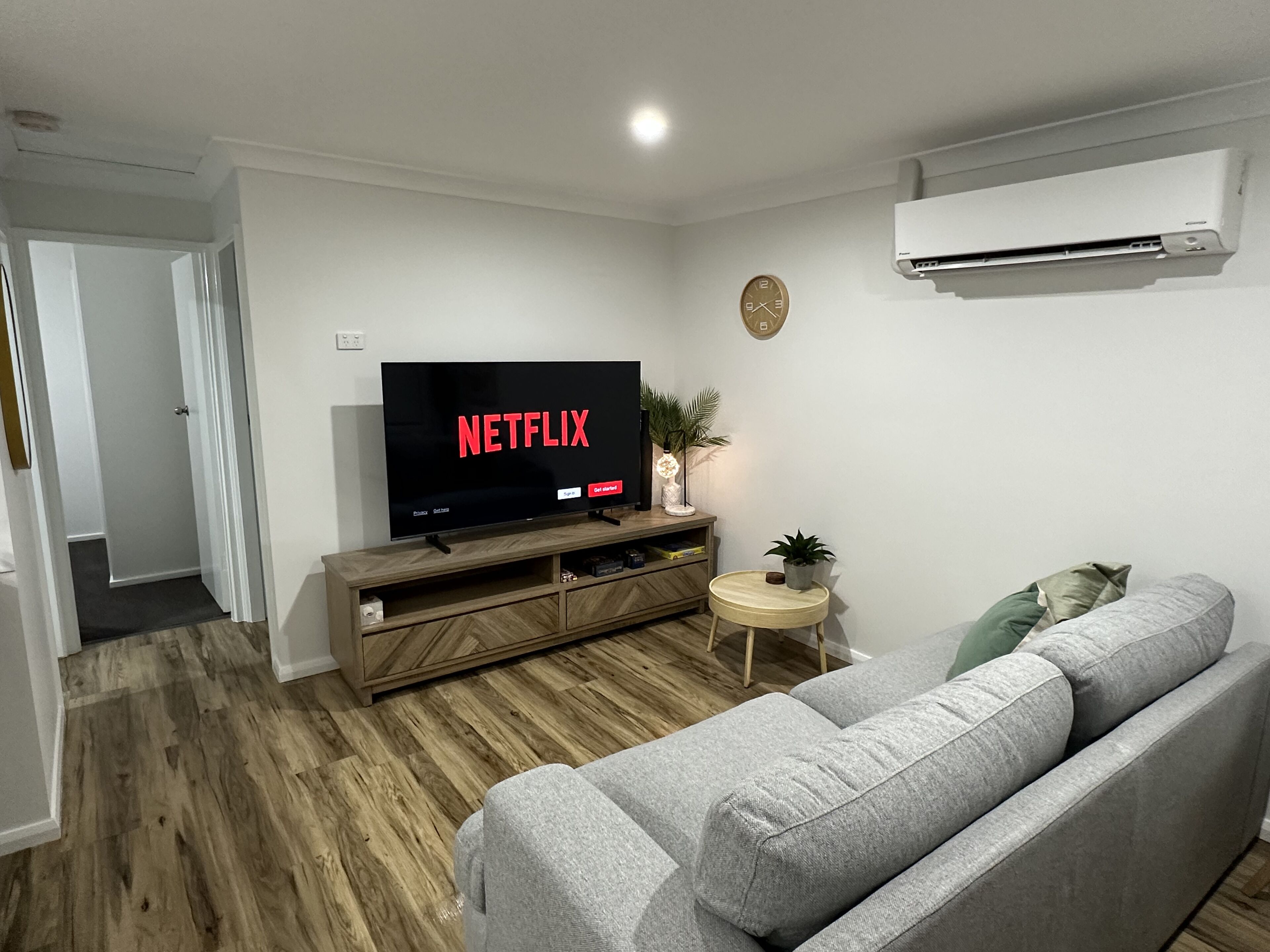 Een woonkamer met een televisie waarop 'NETFLIX' staat, een houten tv-meubel, een grijze bank, een klein rond bijzettafeltje met een plant en een wandairconditioner.