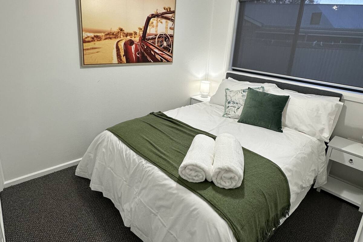 Un lit bien fait, avec du linge de lit blanc et un plaid vert, un ventilateur de plafond, un tableau représentant une voiture accroché au mur et une fenêtre donnant sur un immeuble.