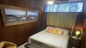 Un dormitorio con cama, un banco, una silla, una mesita de noche, una lámpara y una ventana con vistas a árboles.