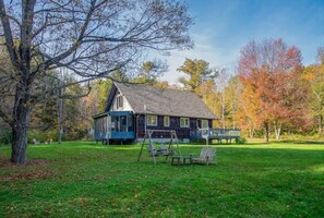 A rustic house with a porch, surrounded by a grassy yard and trees with autumn foliage. - Gorgeous Mountains Views & Private Stream in Secluded Oasis (Gilboa)