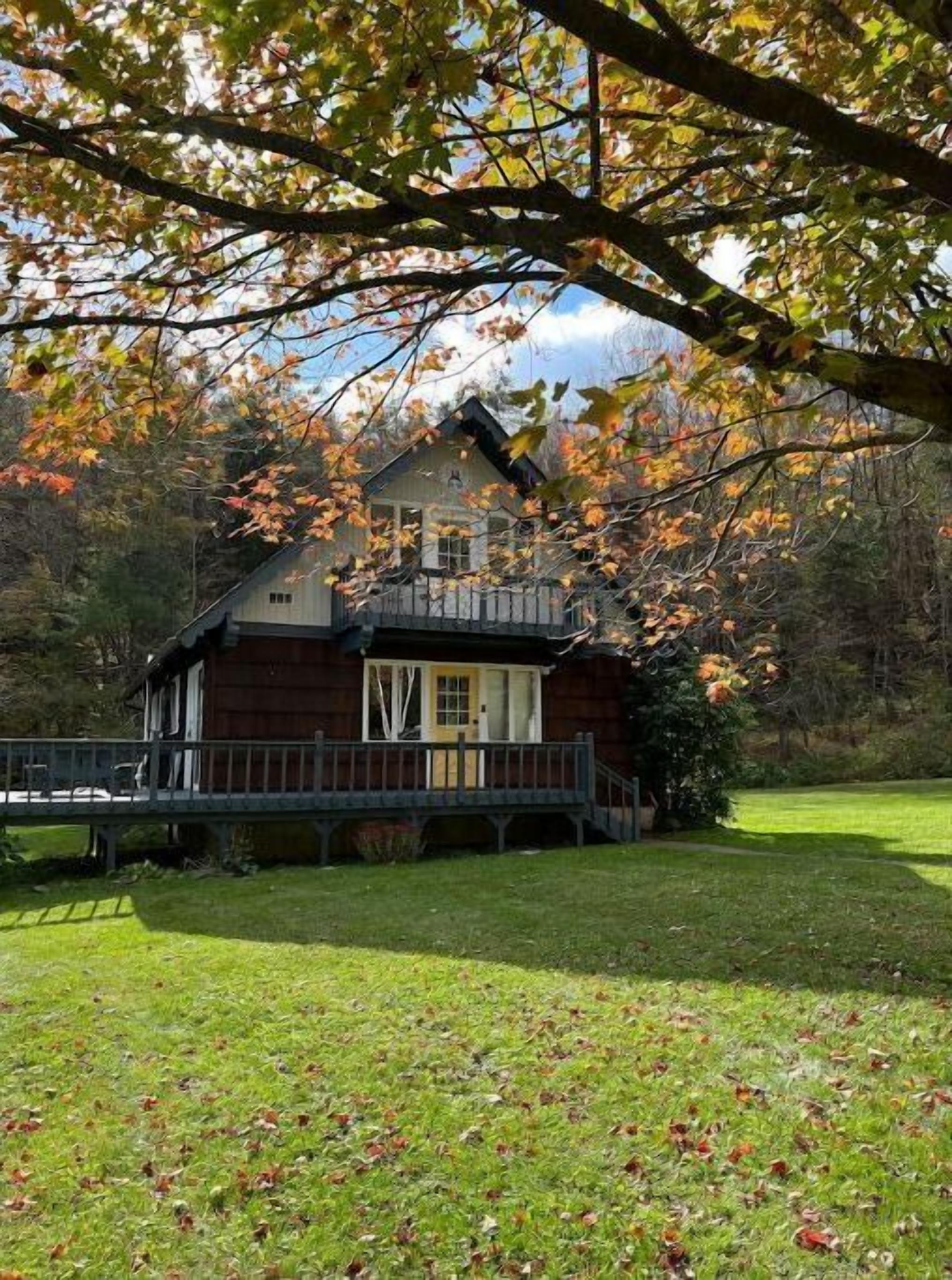 A wooden cabin with a porch, surrounded by a grassy yard and trees with autumn leaves.