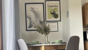 A dining area with a wooden table and two grey chairs, a vase with dried plants, and two framed pictures on the wall.
