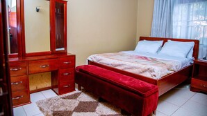 A bedroom with a bed, a wooden dresser, a mirror, and a window with curtains. - Beautiful 2-Bed house in Kampala (Kampala)