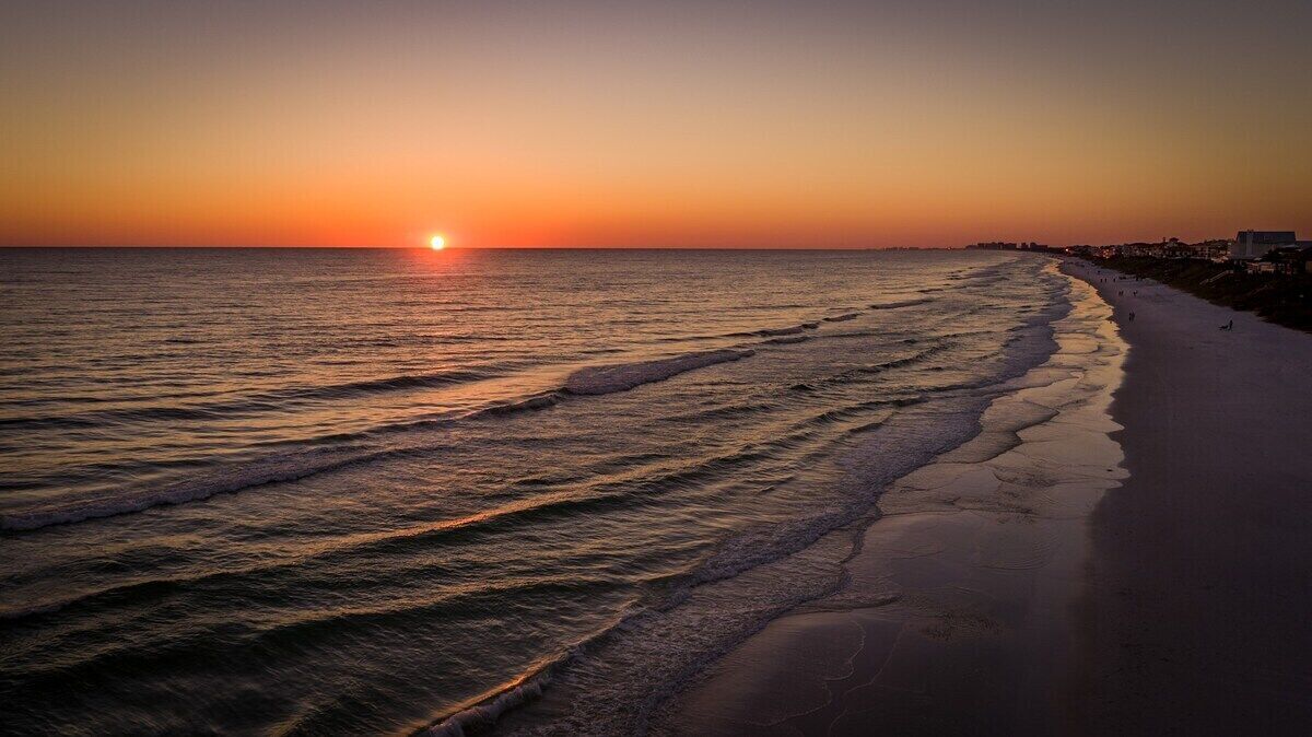 Una playa al atardecer con olas, cielo despejado y una costa lejana.