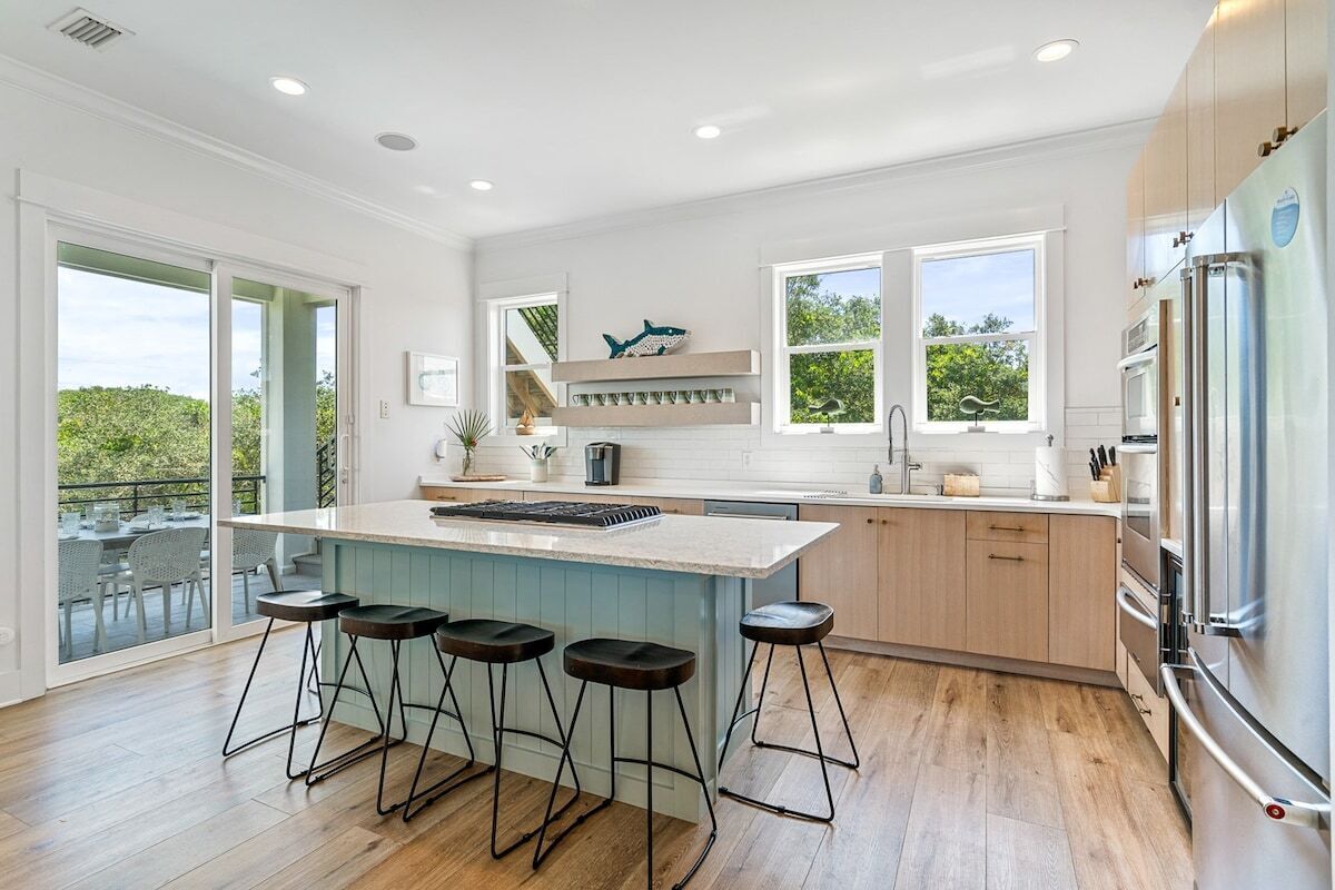 A modern kitchen with a central island, bar stools, a large refrigerator, and a view through the glass doors.