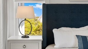 Una camera da letto con un letto, un comodino con una lampada, una finestra vista su del verde e un quadro appeso al muro.
