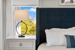 Ein Schlafzimmer mit einem Bett, einem Nachttisch mit Lampe, einem Fenster mit Blick ins Grüne und einem an der Wand hängenden Bilderrahmen.