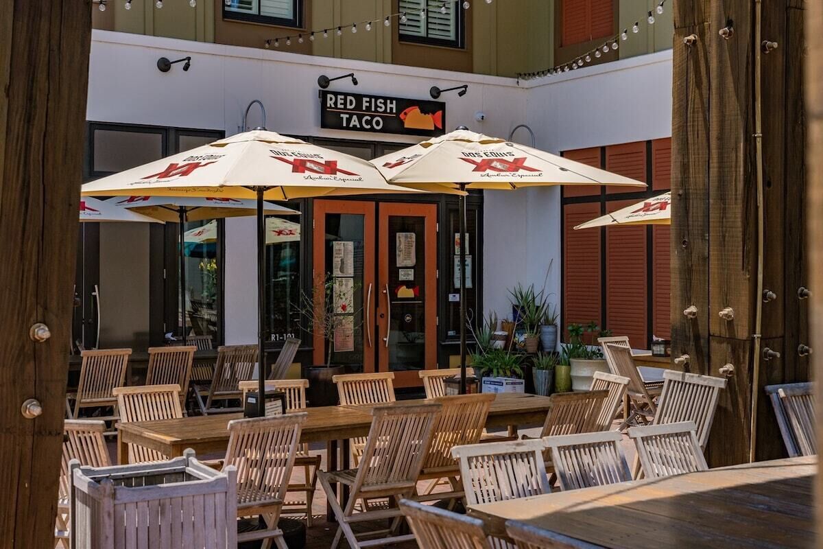 Un'area pranzo all'aperto con tavoli e sedie di legno, coperta da ombrelloni con un logo, e un cartello per il ristorante 'Red Fish Taco'.