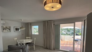 Un salon moderne avec un canapé, une table à manger et des chaises. Il y a une grande fenêtre avec des rideaux, un luminaire au plafond et une vue sur un balcon dont le sol est carrelé.
