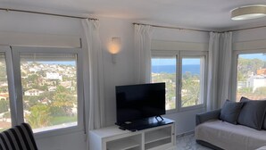 Un salon moderne avec un téléviseur posé sur un meuble blanc, un fauteuil à rayures, un canapé gris et de grandes fenêtres offrant une vue sur la ville et la mer.