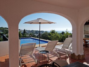 A pool area with two lounge chairs and an umbrella, overlooking a scenic view of the sea and a town. - Meerblick, Apartment in Villa mit Terrasse, Garten, Pool und Kostenlosen Wlan (Benissa)