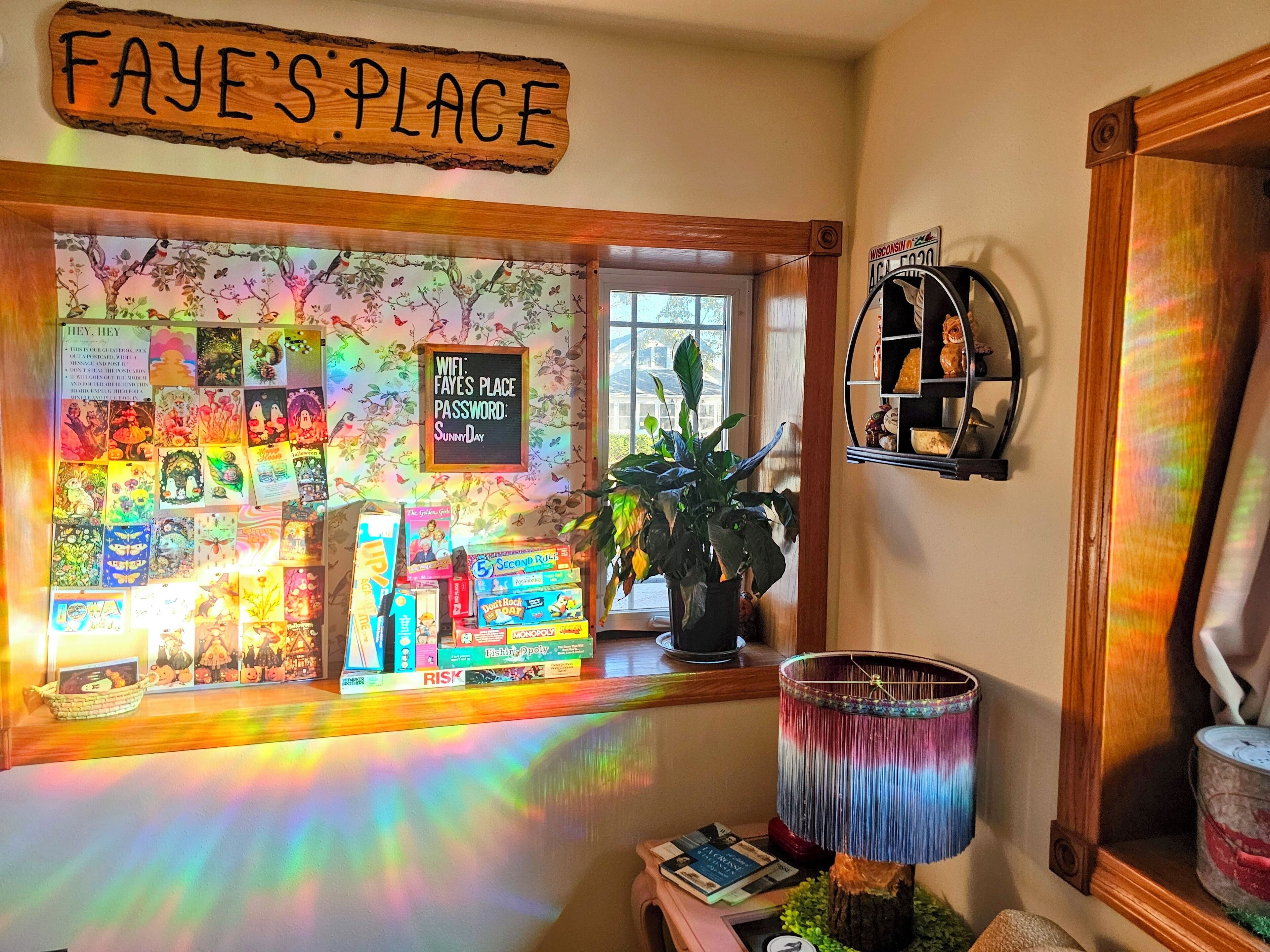Una habitación acogedora con un letrero que dice "Faye's Place", una ventana con ilustraciones coloridas, una lámpara y una estantería con diversos objetos.