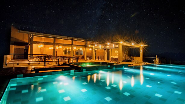 Piscina iluminada à noite sob um céu estrelado, um edifício com janelas iluminadas e um deque bem iluminado.