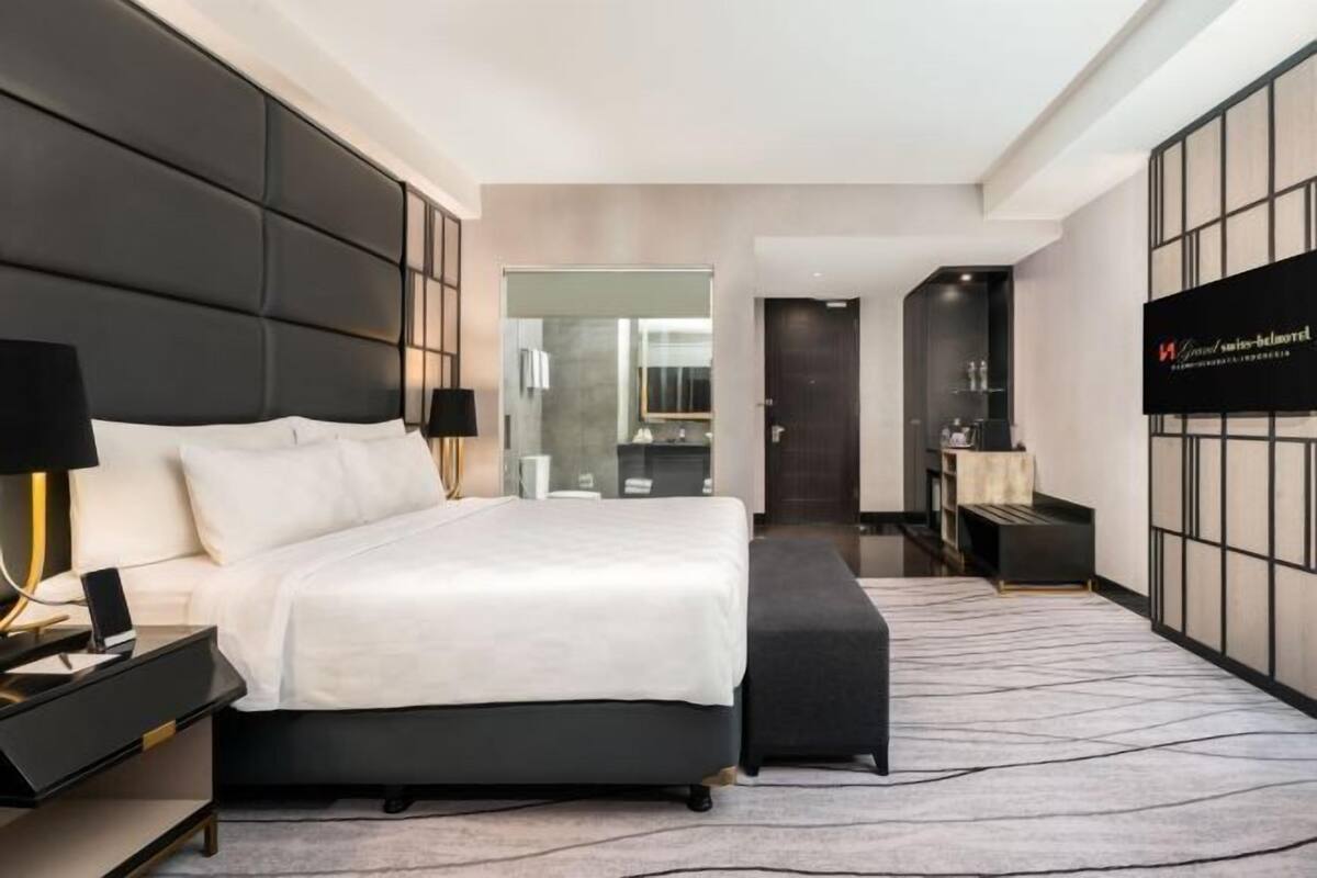Habitación de hotel moderna con una cama grande, mesitas de noche, un televisor y un baño visible a través de una puerta de vidrio.