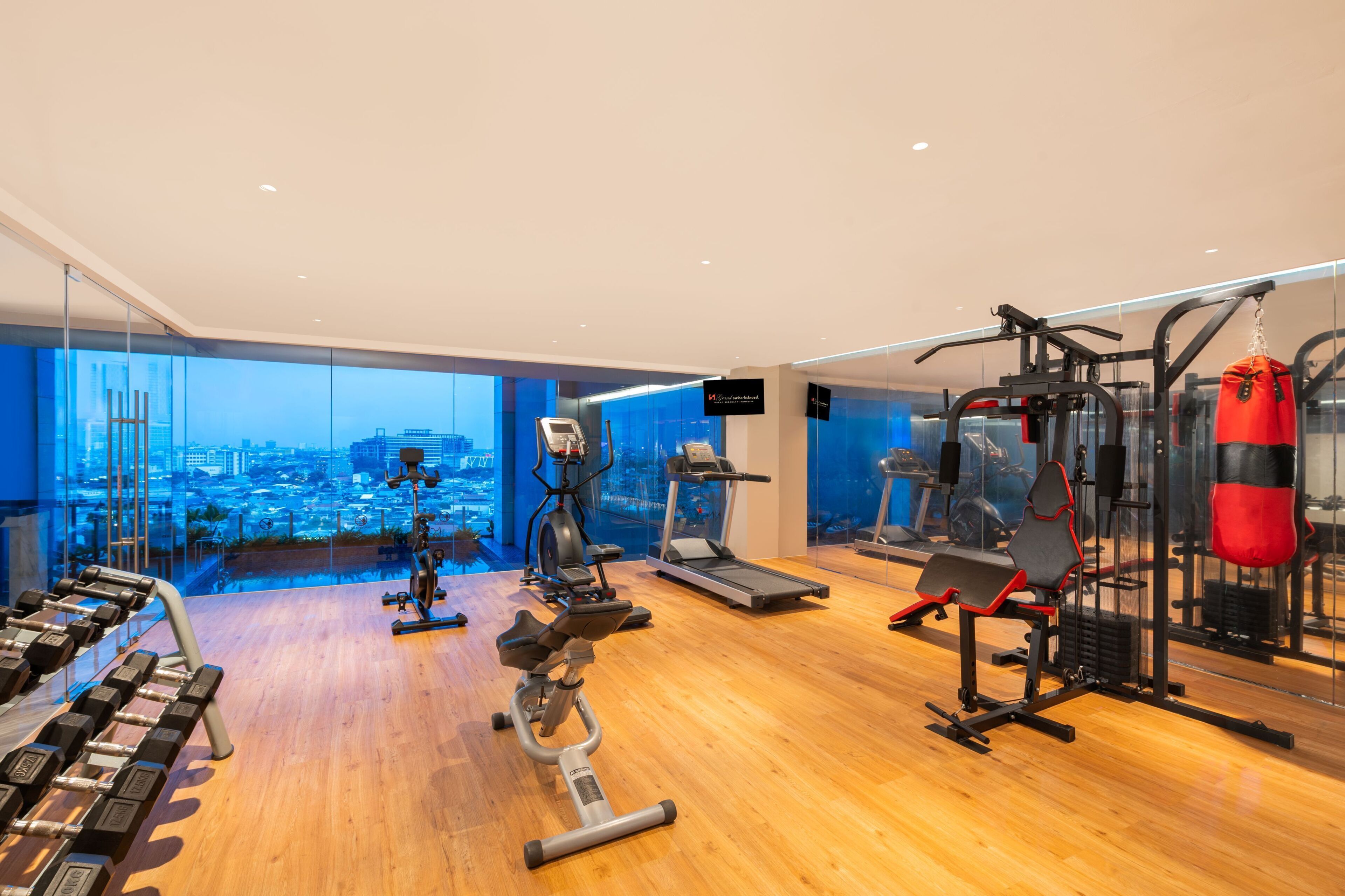Academia moderna com equipamentos de cardio, aparelhos de musculação e saco de pancada, com grandes janelas e vista para a cidade.