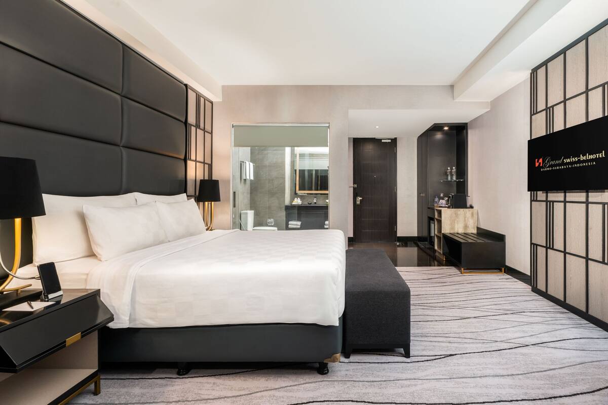 Habitación de hotel moderna con una cama grande, mesitas de noche, un televisor y un baño visible a través de una puerta de vidrio.