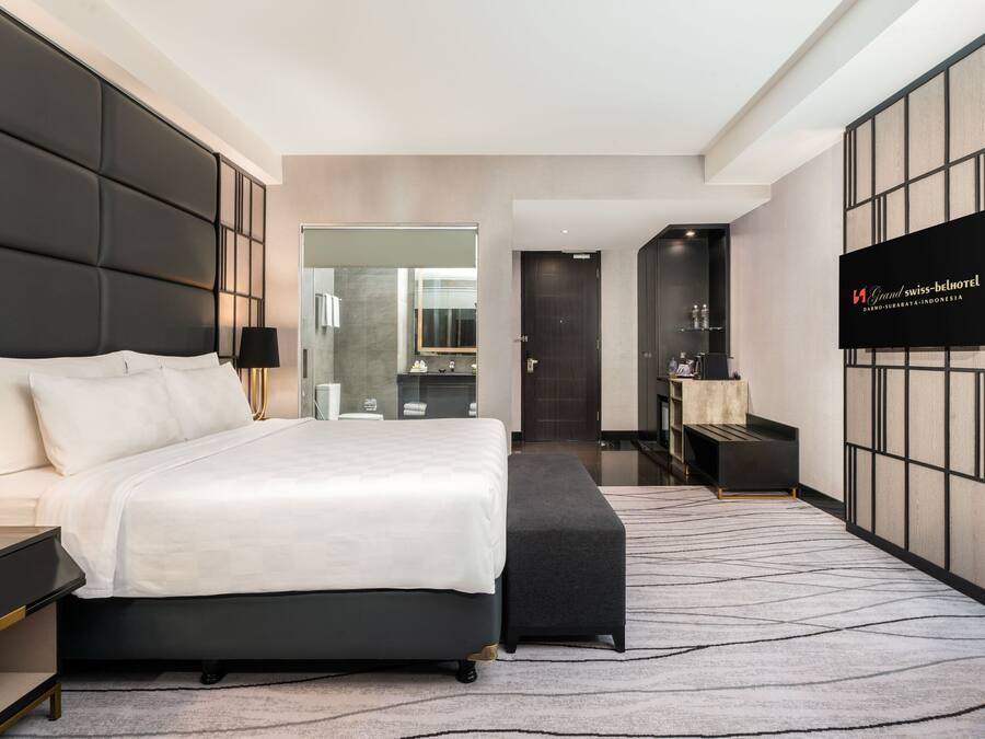 A modern hotel room with a large bed, bedside tables, a TV, and a bathroom visible through a glass door.