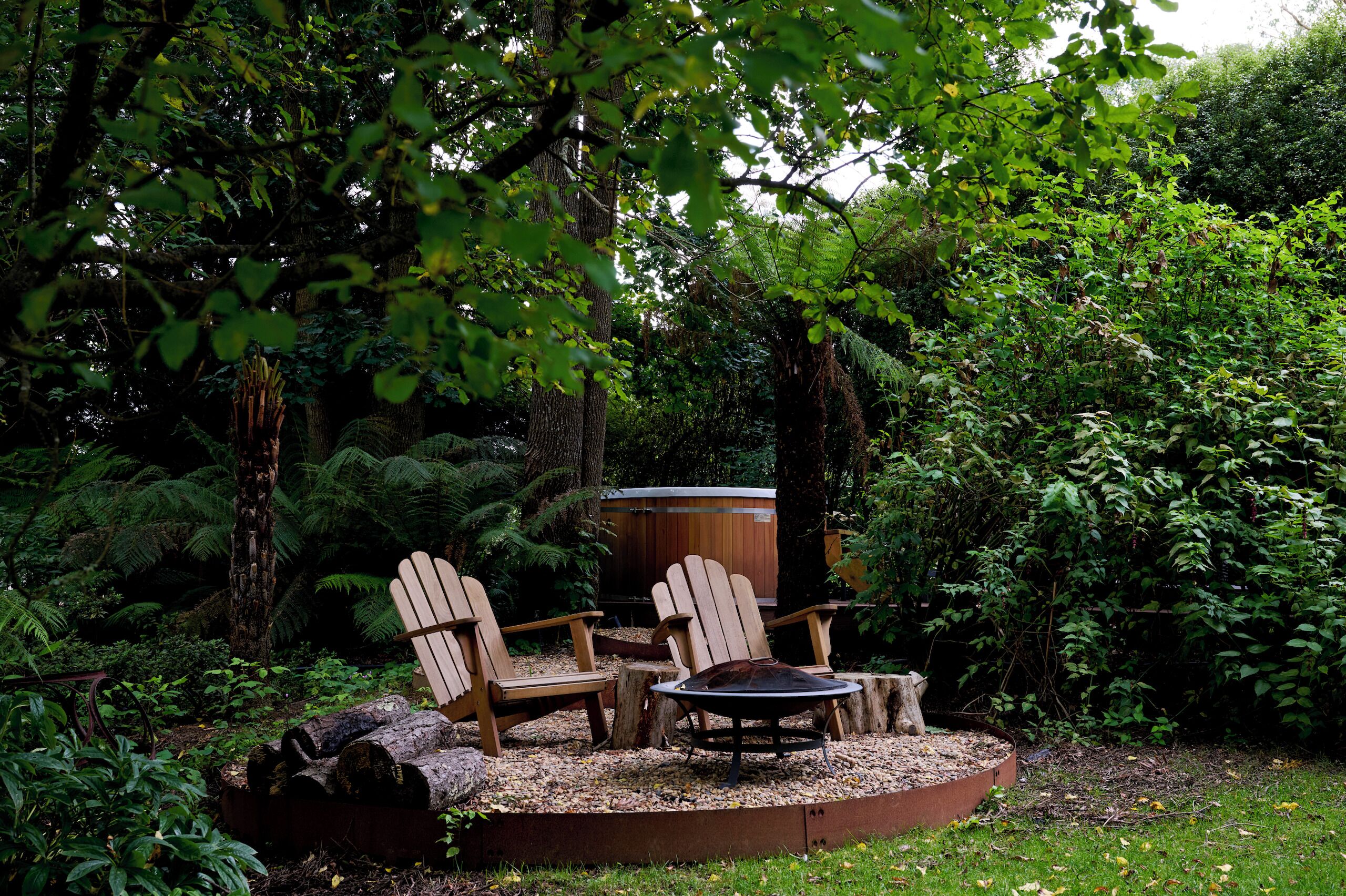 Een houten terras met Adirondack-stoelen en een ronde tafel, omringd door boomstammen, gelegen in een weelderige tuin met dichte begroeiing en een houten constructie op de achtergrond.