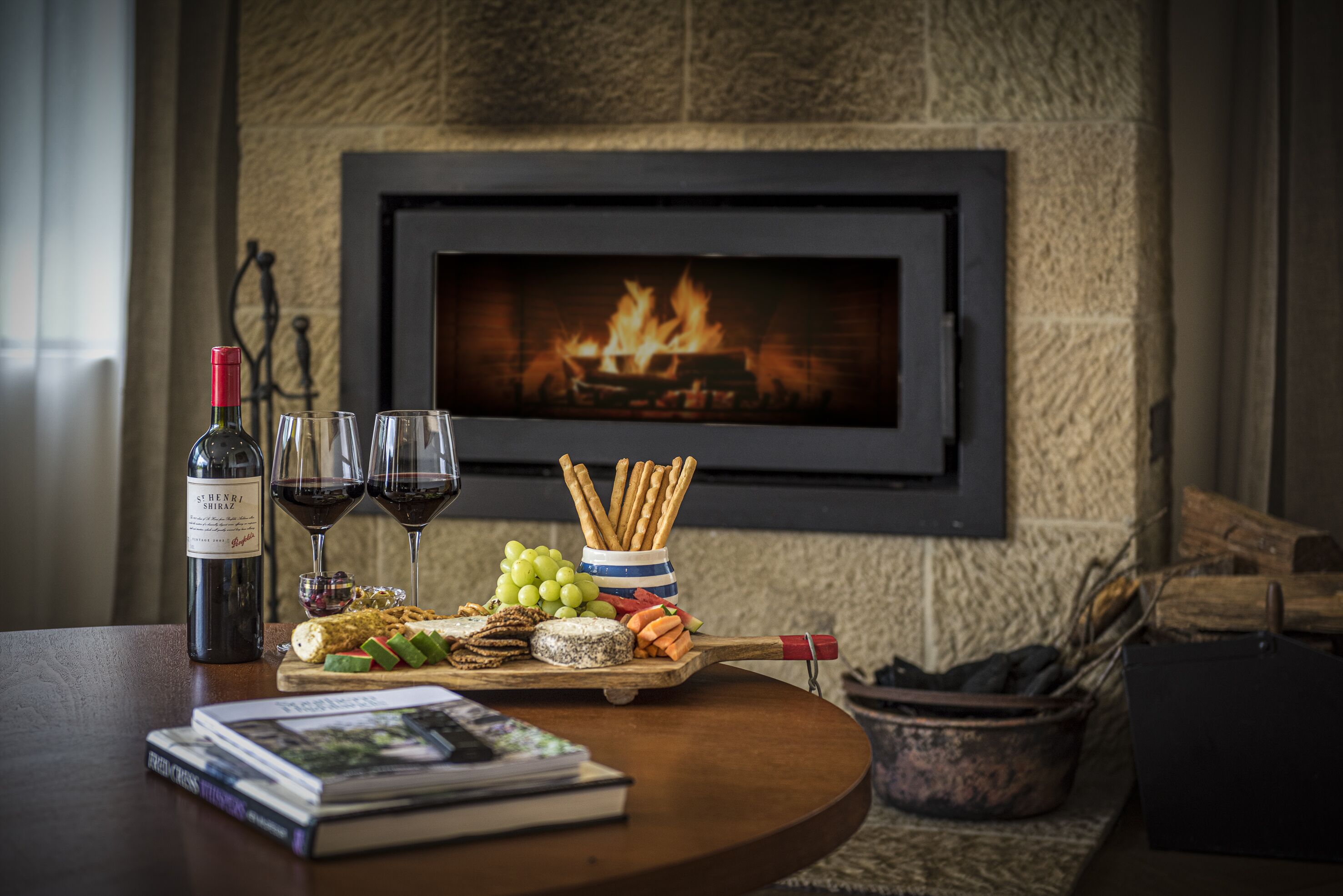 A table with a bottle of wine, glasses, a bowl of snacks, and a book. A fireplace with a fire is in the background.