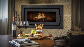 A table with a bottle of wine, glasses, a bowl of snacks, and a book. A fireplace with a fire is in the background.