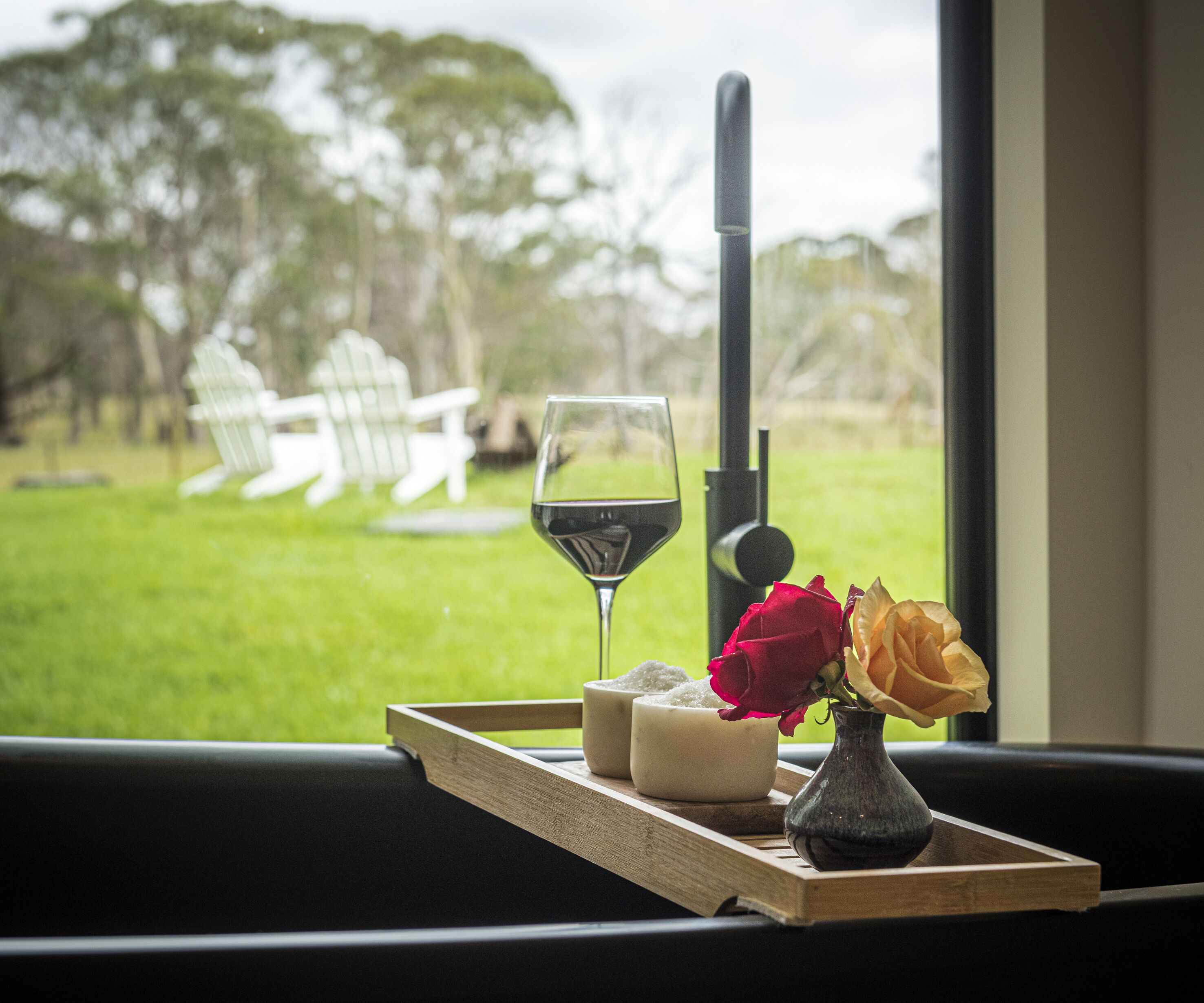 A tray with a glass of red wine, two candles, and a vase with flowers on a window sill.