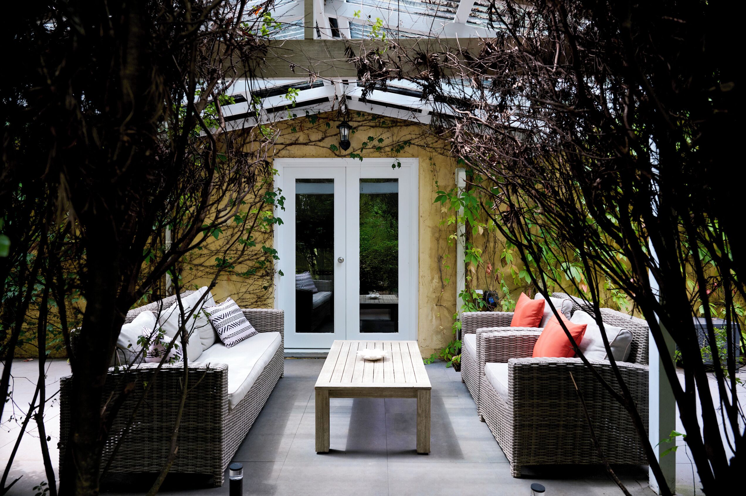 A cozy outdoor seating area with wicker furniture and a central table, surrounded by greenery and a stone wall.