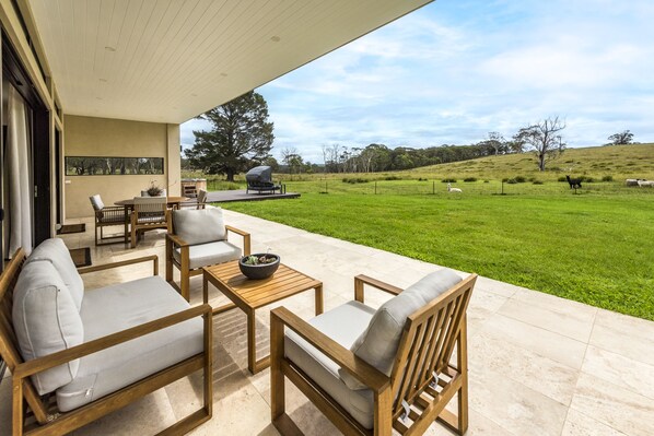 A patio with outdoor furniture, a dining table, and a view of a grassy field with trees and a hill in the background. - The Vale Penrose (Penrose)