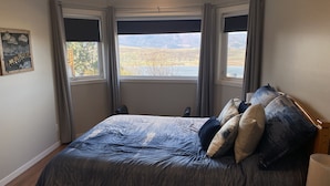 Un dormitorio con una cama grande, dos ventanas con cortinas, vista a un lago y montañas, y un cuadro en la pared.