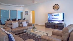 A modern living room with a glass coffee table, leather sofas, a flat-screen TV, and a kitchen area visible through an open door. - 6 Bedroom Tropical Pool Villa - V6 (Pattaya)