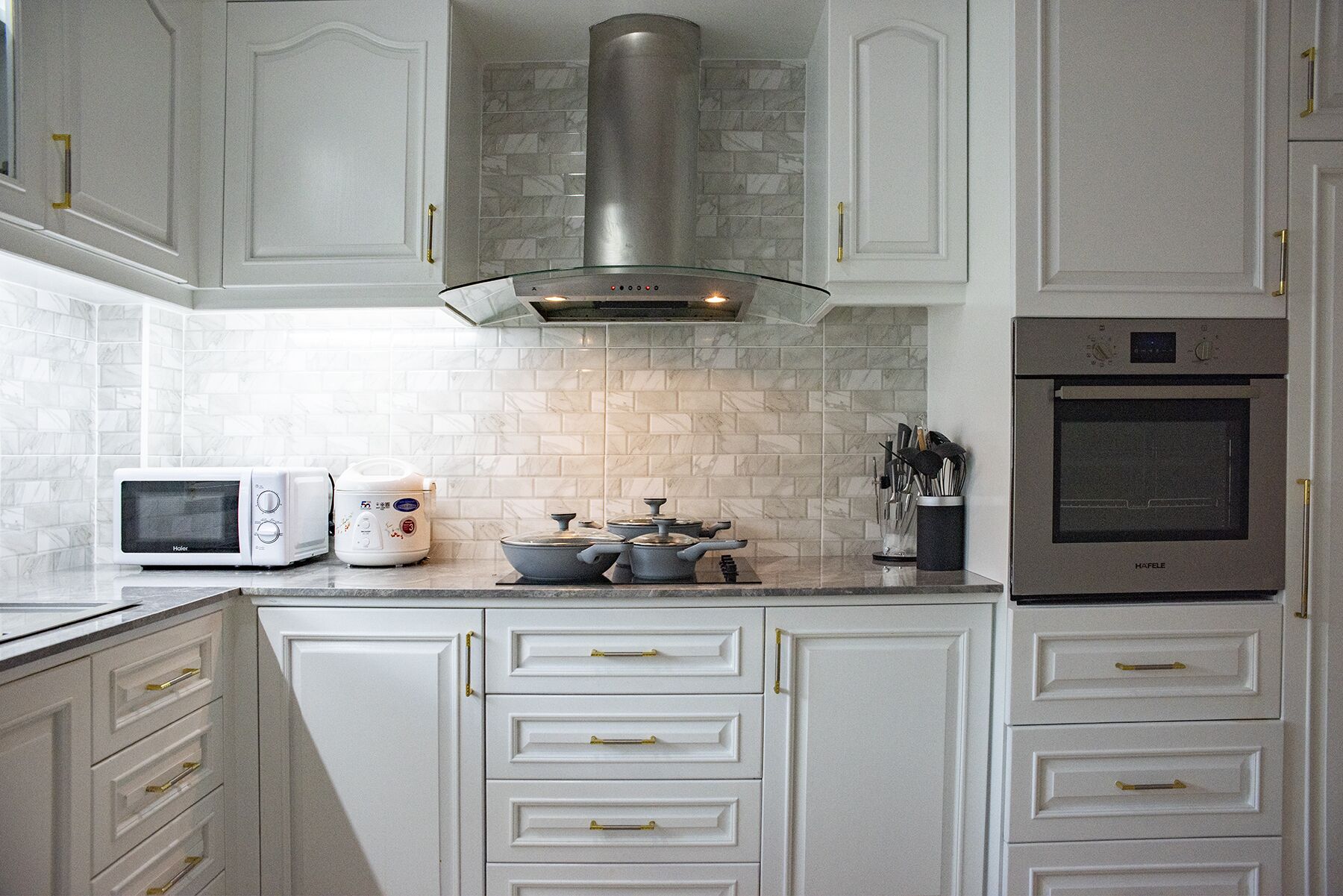A modern kitchen with white cabinetry, a stainless steel range hood, a microwave, a rice cooker, and an oven.