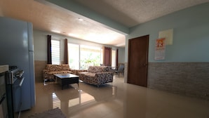 A spacious living area with a sofa, coffee table, and a dining table. A refrigerator is visible on the left, and a door is on the right. - Taina - Grand Appartement Avec Terrasse Avec Accès mer (AVERA - TAPUTAPUATEA)