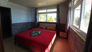 A bedroom with a bed, a nightstand, a window with curtains, and a view of palm trees outside. - Taina - Grand Appartement Avec Terrasse Avec Accès mer (AVERA - TAPUTAPUATEA)
