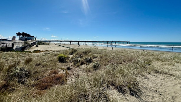Ett kustlandskap med en pir som sträcker sig ut i havet, sanddyner och en byggnad med modern design.