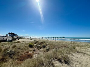 A coastal landscape with a pier extending into the sea, sandy dunes, and a building with a modern design.