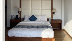 Habitación de hotel ordenada con una cama grande, cabecero de madera, dos mesitas de noche con lámparas y una mesita auxiliar con lámpara.