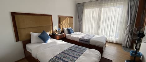 Habitación de hotel con dos camas, una mesita de noche con lámpara, un ventilador y una ventana con cortinas.