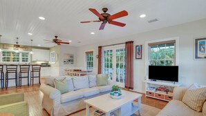 A spacious living room with a white sofa, a coffee table, and a TV. There is a kitchen area with white cabinets and a dining table with chairs. - Relaxing Getaway w/ Private Pool (Tybee Island)