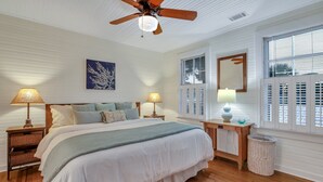 A bedroom with a large bed, two bedside tables with lamps, a ceiling fan, a window with shutters, and a wall-mounted mirror. - Relaxing Getaway w/ Private Pool (Tybee Island)