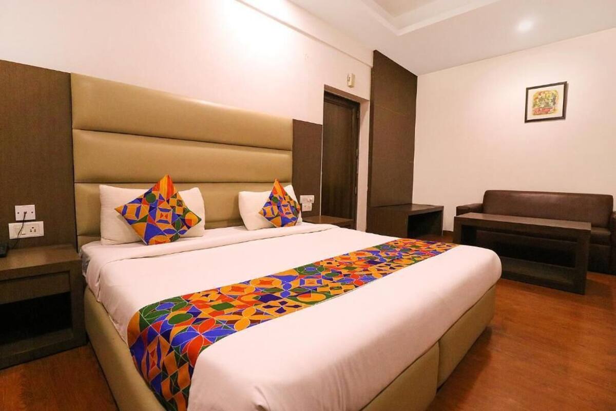 A hotel room with a large bed, two pillows, a colorful blanket, a wooden nightstand, a brown sofa, and a framed picture on the wall.
