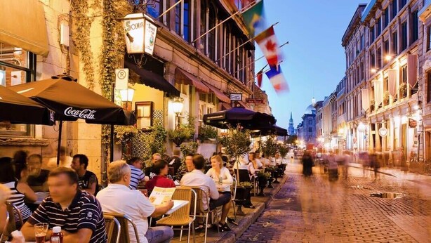 Eine Straße mit Straßencafés und Sitzgelegenheiten, ein Kopfsteinpflasterweg und Gebäude mit Fahnen.